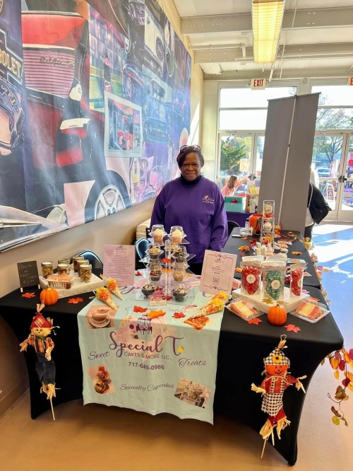 A vendor stands behind a decorated table with baked goods and crafts, featuring fall-themed decorations and jars.