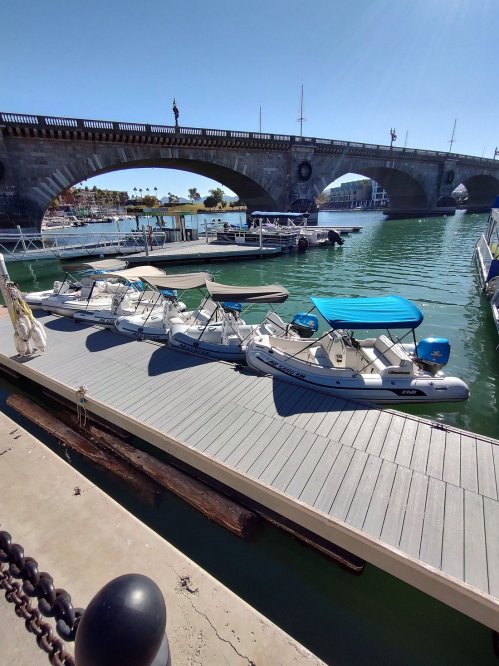 A marina with several boats docked on a wooden pier under a stone bridge on a sunny day.