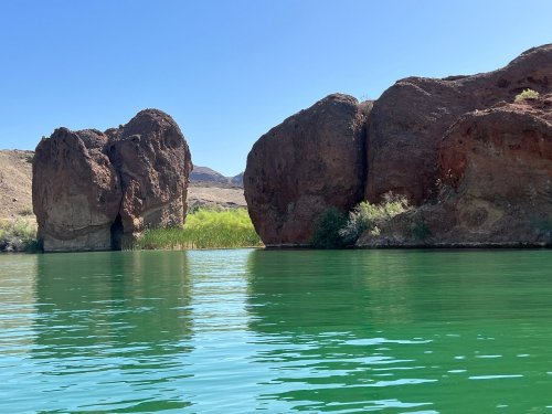 A serene lake scene with green water, flanked by large, rocky formations and lush vegetation under a clear blue sky.