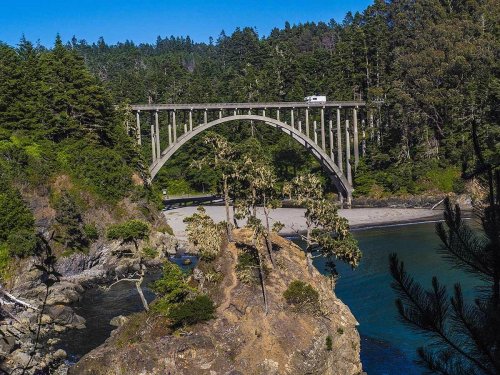 A scenic view of a concrete arch bridge over a river, surrounded by lush greenery and rocky shores.