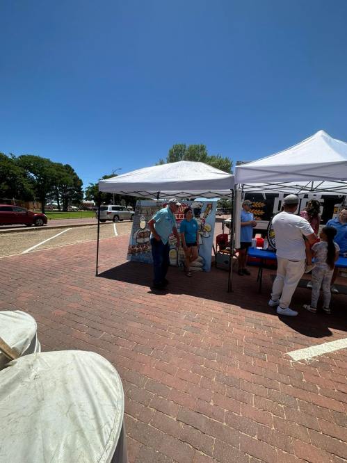 A sunny outdoor market scene with tents, people mingling, and a backdrop featuring a colorful mural.