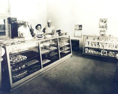 A vintage bakery interior with three bakers behind a glass display filled with various baked goods.