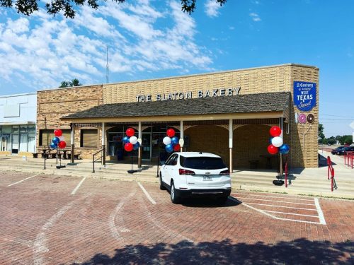 Exterior of Slaton Bakery with red, white, and blue balloons, a parked white car, and a clear blue sky.