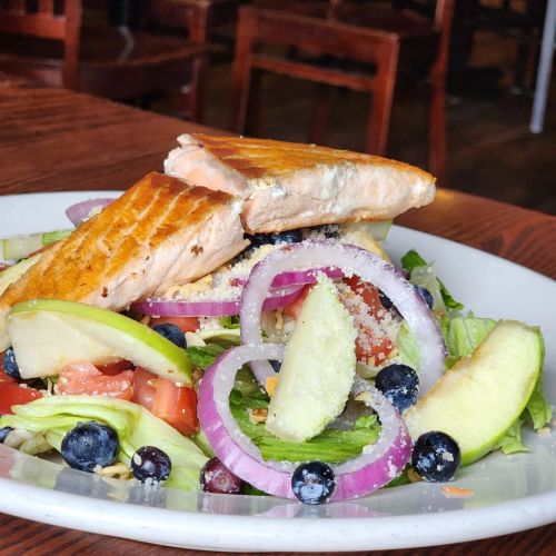 A colorful salad topped with grilled salmon, apples, blueberries, onions, and a sprinkle of cheese on a white plate.