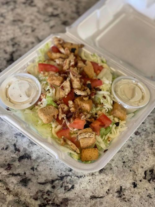 A salad in a takeout container with lettuce, tomatoes, grilled chicken, croutons, and two dressing cups on a granite surface.