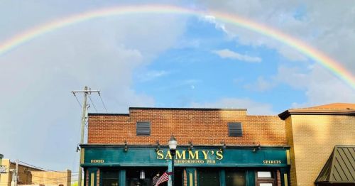 A vibrant rainbow arches over Sammy's Neighborhood Pub, a brick building with a sign for food and spirits.