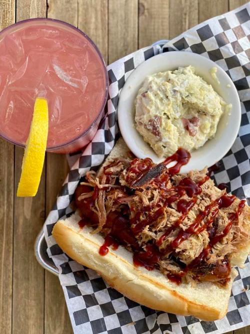 A pulled pork sandwich with barbecue sauce, a side of potato salad, and a pink drink with a lemon slice.