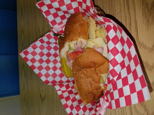 A basket with two sandwiches, pickles, and potato chips, lined with red and white checkered paper.