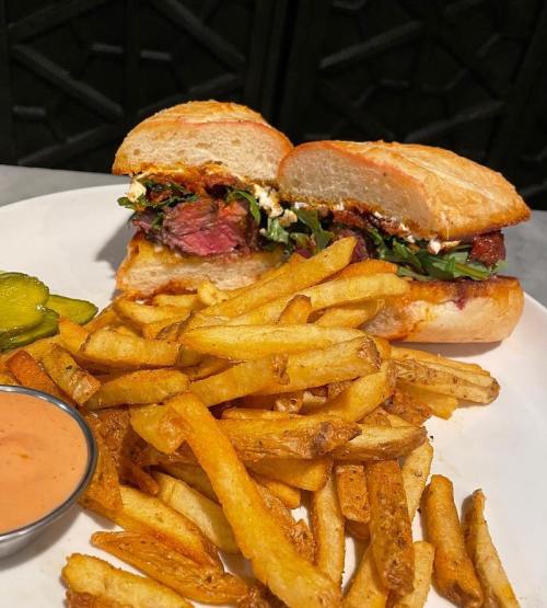 A plate featuring a sliced sandwich with meat and greens, accompanied by crispy fries and a side of dipping sauce.