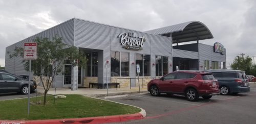 A modern restaurant building with a metal exterior, featuring large windows and parked cars in front.