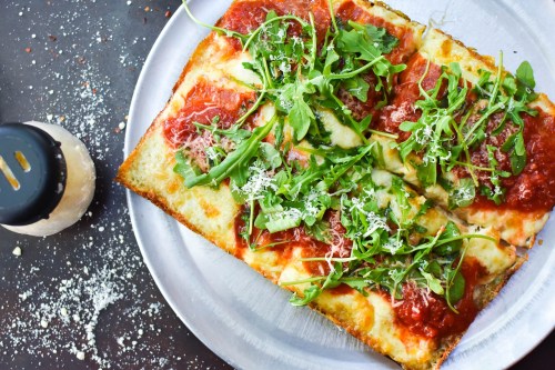 A rectangular pizza topped with marinara sauce, cheese, and fresh arugula, served on a metal plate with grated cheese nearby.