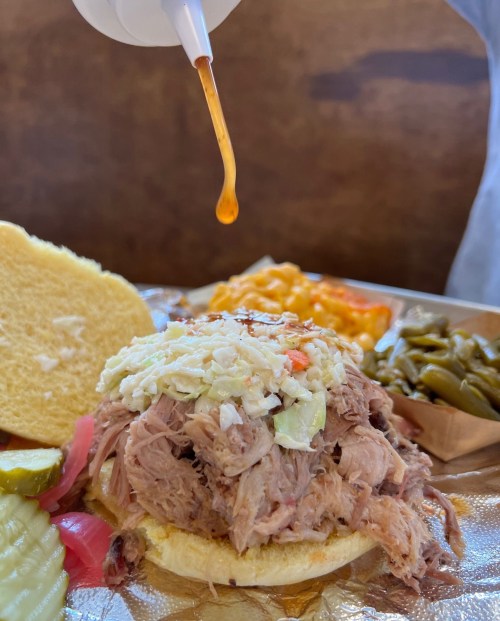 A close-up of a pulled pork sandwich topped with coleslaw, served with pickles, macaroni, and green beans.