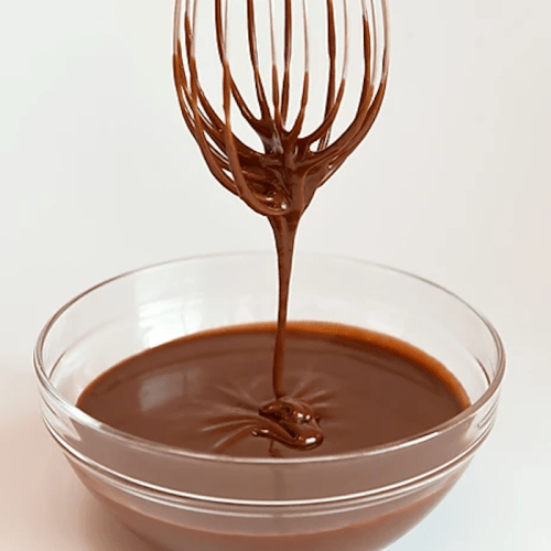 A whisk dripping with rich chocolate batter above a bowl of smooth chocolate.