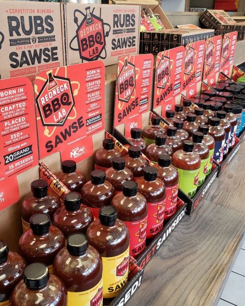 A display of various BBQ sauces and rubs in bottles, showcasing different flavors and brands on a wooden shelf.