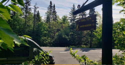 A trail sign for Sawyer Mountain, 1.0 miles, with a view of a road and trees in the background.