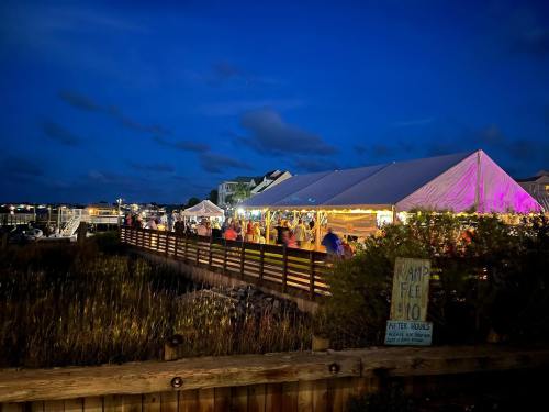 A lively outdoor event under a large tent at dusk, with colorful lights and a waterfront view.