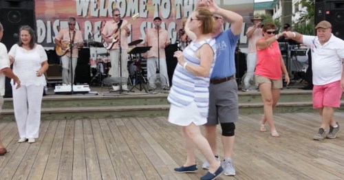 A lively outdoor dance scene with people enjoying music on a wooden stage, smiling and dancing together.