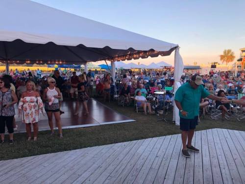 A lively outdoor event with a large crowd under tents, featuring people dancing and enjoying the atmosphere.