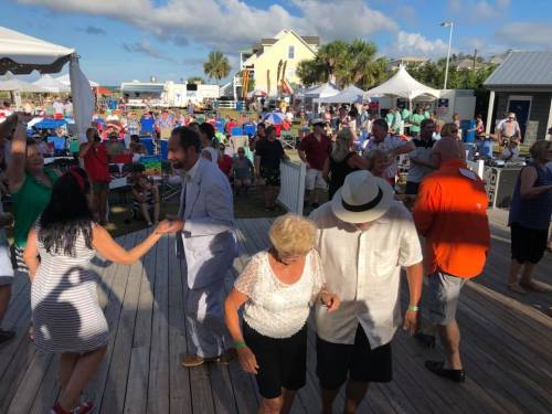 A lively outdoor event with people dancing, enjoying music, and socializing under a sunny sky. Tents and chairs in the background.