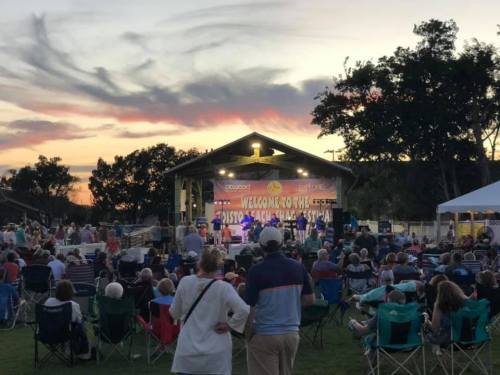 A crowd gathers at an outdoor concert during sunset, with a stage and colorful sky in the background.