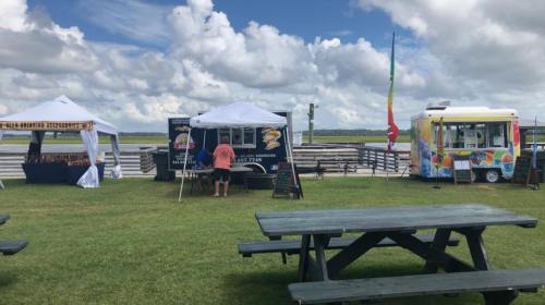 A grassy area with food trucks and tents under a cloudy sky, featuring picnic tables in the foreground.