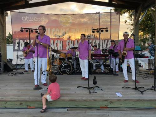 A band in pink shirts performs on stage while a child sits on the floor, enjoying the music at a festival.