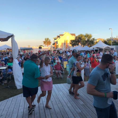 A lively outdoor event with a crowd enjoying music, food, and activities under tents and sunny skies.