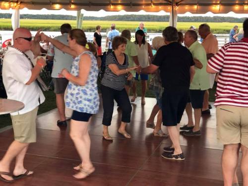 A lively outdoor dance scene with people enjoying music under a tent by a waterfront.