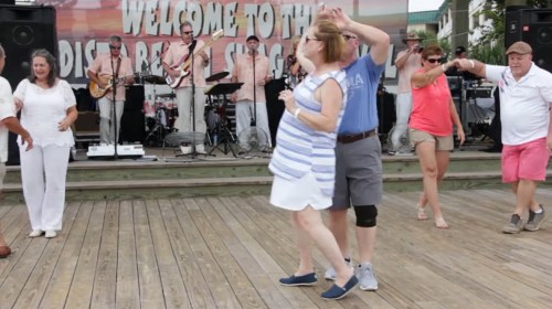 People dancing on a wooden stage with a live band in the background, enjoying a festive atmosphere.
