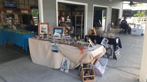 A craft market scene with tables displaying handmade items, including jewelry and framed art, in a covered area.