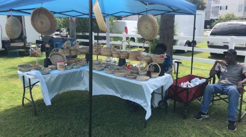A vendor tent displays various woven baskets, with two people seated nearby in a grassy area.