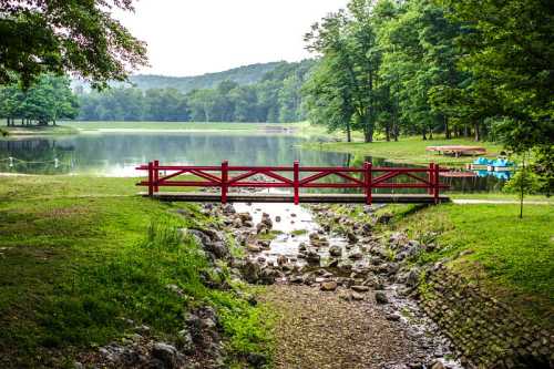 A red wooden bridge spans a small stream leading to a serene lake surrounded by lush green trees.