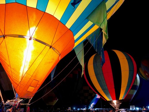 Colorful hot air balloons illuminated at night, with flames visible in one balloon and vibrant patterns on others.