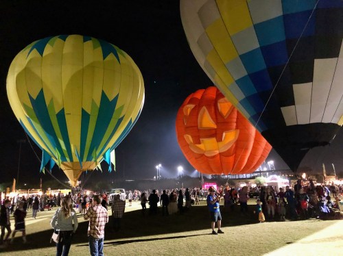Colorful hot air balloons, including a pumpkin-shaped one, illuminate a night festival with crowds enjoying the event.