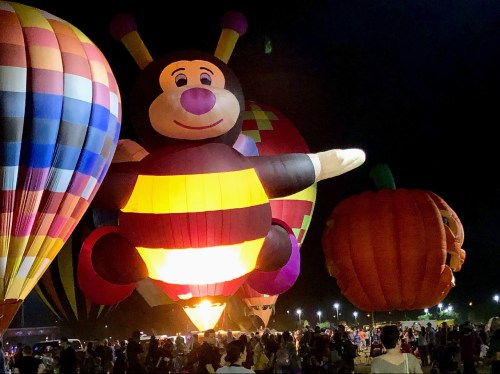 Colorful hot air balloons, including a large bee and a pumpkin, illuminate the night sky with a crowd below.