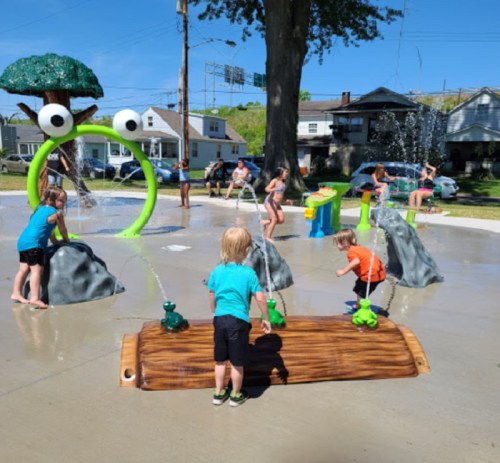 Children play in a splash pad with water features, rocks, and colorful structures on a sunny day.