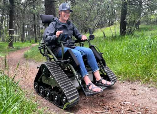 A person in a wheelchair with tracks smiles while navigating a forest path surrounded by greenery.