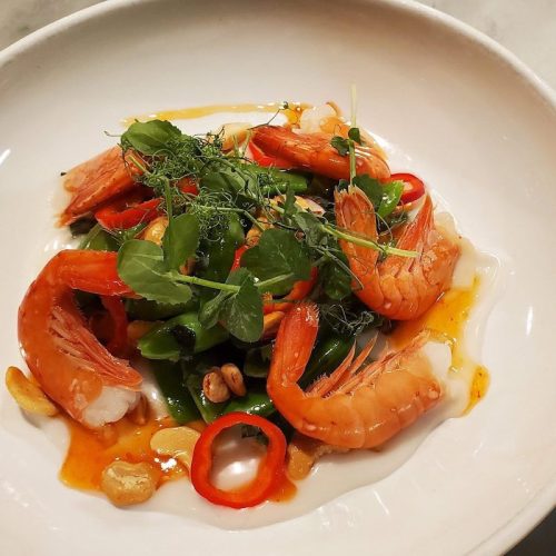 A bowl of shrimp salad with green beans, red chili, herbs, and peanuts drizzled with a spicy sauce.