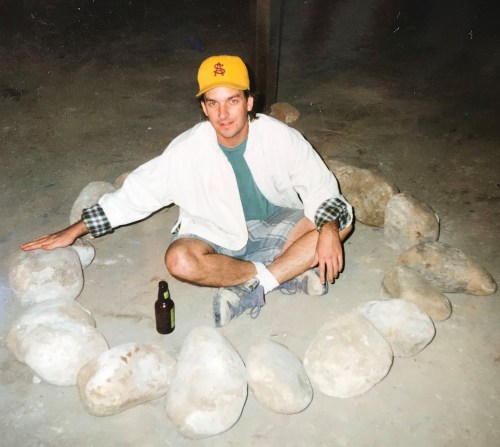 A man sits cross-legged in a circle of rocks, wearing a yellow cap and holding a bottle, in a dimly lit space.