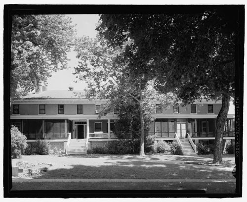 Historic two-story house with large porch, surrounded by trees and a grassy area, in black and white.