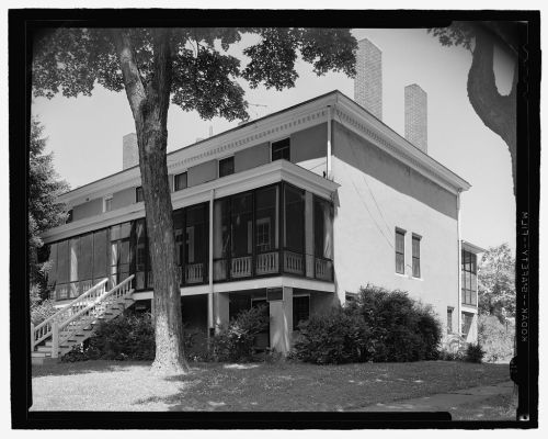 Historic two-story building with a screened porch, surrounded by trees and greenery. Black and white photograph.