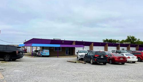 A low-rise building with a purple and blue roof, surrounded by a gravel parking lot filled with various cars.