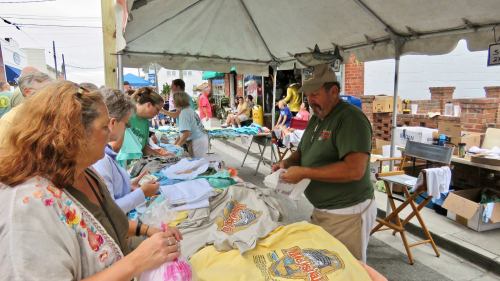 A vendor sells T-shirts at a market, while customers browse and interact under a tent.