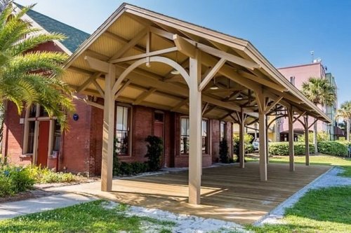 A wooden pavilion with a sloped roof stands in front of a brick building, surrounded by greenery and palm trees.