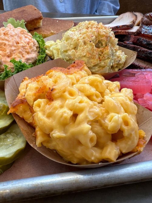 A tray of comfort food featuring creamy macaroni and cheese, pickles, and various salads.