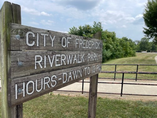 Wooden sign for Riverwalk Park in Frederick, listing hours from dawn to 10 PM, with a path and greenery in the background.