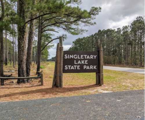 Sign for Singletary Lake State Park, surrounded by tall trees and a gravel road. Cloudy sky in the background.