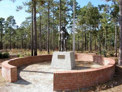 A bronze statue of a soldier stands on a stone pedestal, surrounded by a circular brick seating area in a wooded area.