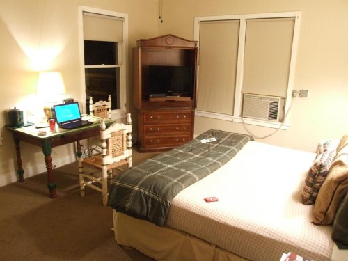Cozy bedroom with a bed, desk, chair, TV, and air conditioner, featuring warm lighting and simple decor.