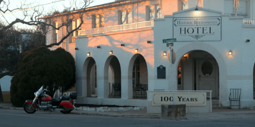 Historic Rocksprings Hotel with a motorcycle parked outside, featuring arched windows and a sign celebrating 100 years.